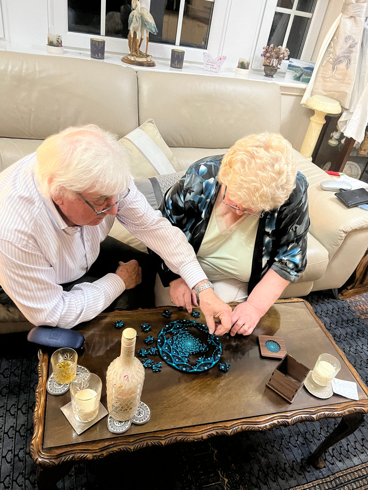 An overhead view of two elderly people gathered around a wooden table in a living room, working together to assemble or decorate a small blue decorative object. The room contains a beige sofa, a patterned rug, and various home decor items.
