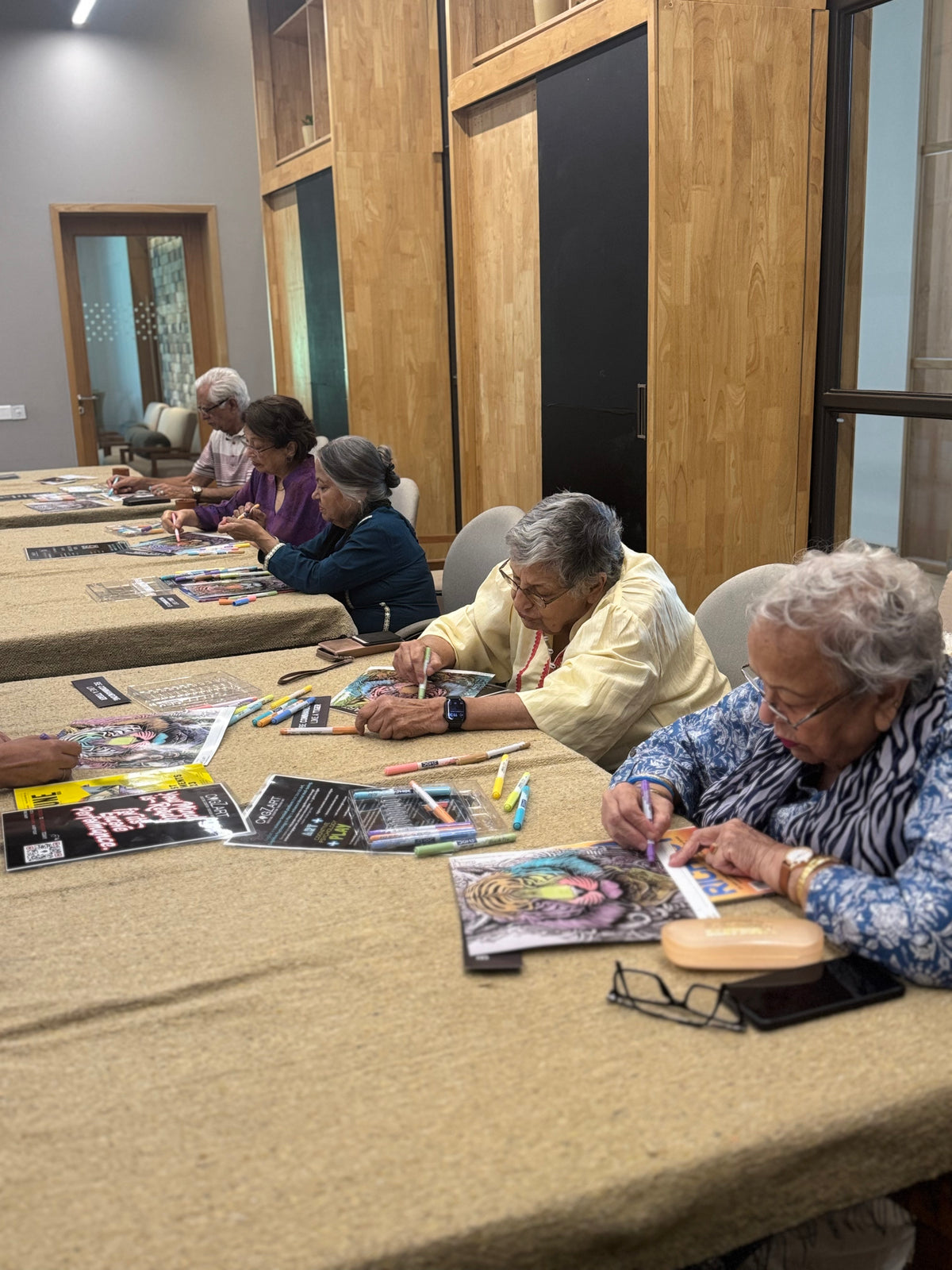 Several people seated along a long table in a room with wood-paneled walls, working on colorful art or craft projects with papers, pencils, and small tools.