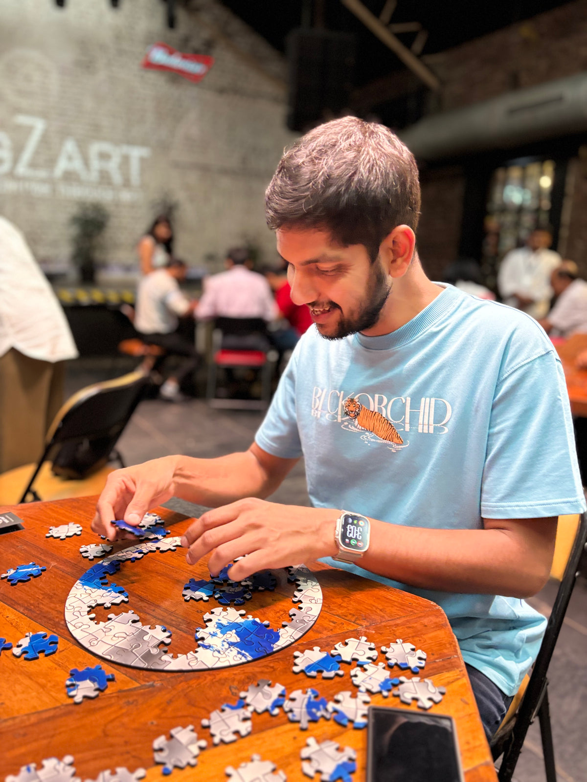 A man wearing a light blue T-shirt with a 'BIOARCH' logo sits at a wooden table, assembling a circular jigsaw puzzle with blue, white, and silver pieces in a busy indoor setting.
