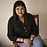 A woman with long dark hair wearing a black top and a brown leather chair against a dark background.